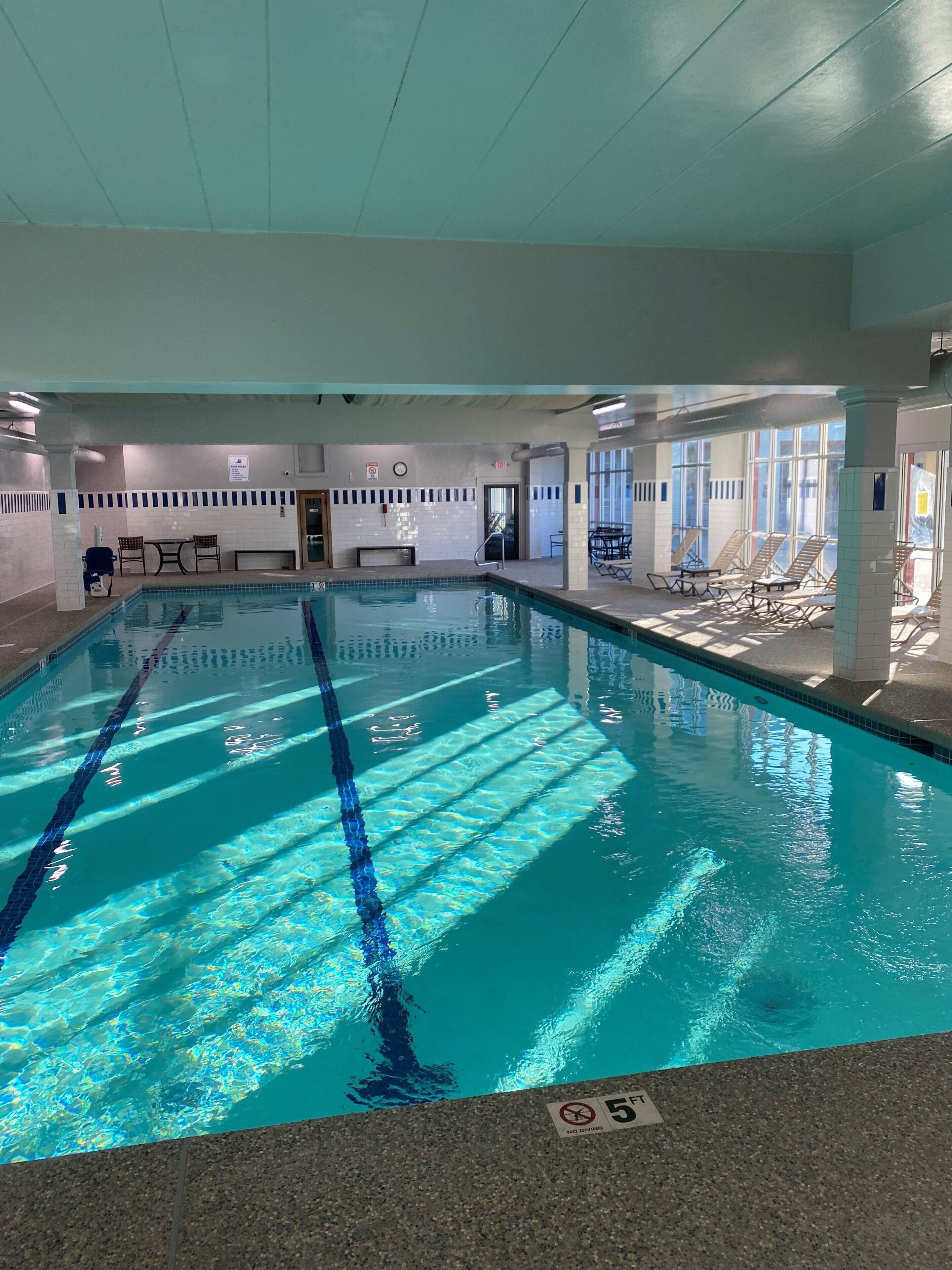 An indoor swimming pool with clear water, marked lanes, and sunlit areas with lounge chairs.