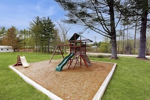 A playground with a slide and climbing structure, surrounded by grass and trees.