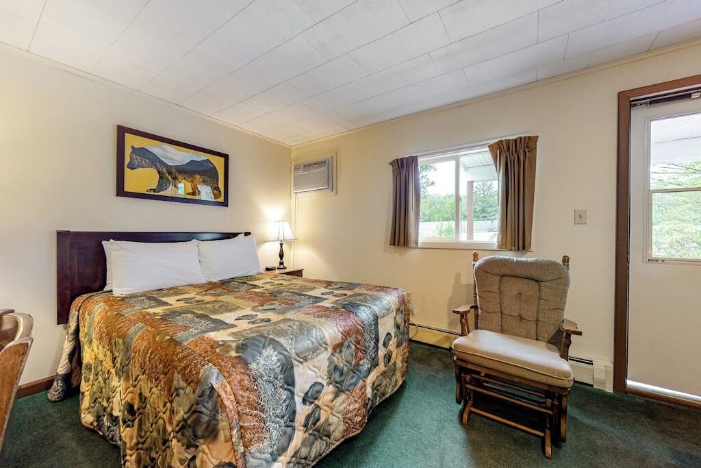 Hotelkamer met een bed, een stoel, een raam met gordijnen, een lamp en een schilderij aan de muur.