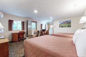 A hotel room with two beds, a desk, a chair, a small kitchenette, and a window with curtains.