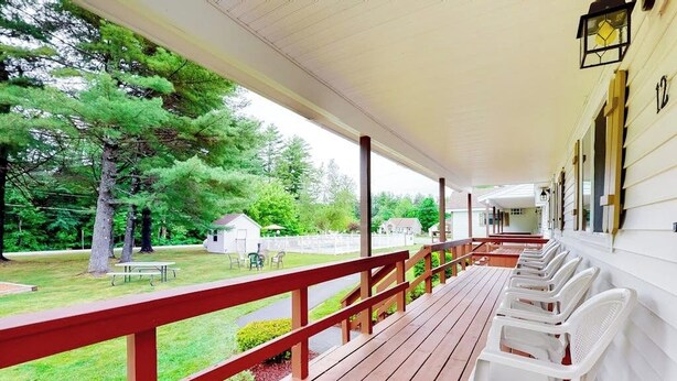 Une véranda couverte avec une terrasse en bois, des chaises en plastique blanches et une vue sur un espace verdoyant agrémenté d’arbres et d’un petit bâtiment.