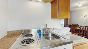 Una cocina con estufa de acero inoxidable, un lavamanos, armarios de madera y un comedor con sillas.