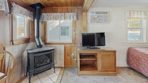 A cozy room with a wood-burning stove, a television on a wooden stand, and a bed with a pink quilt. - Eclectic Studio Cabin w/ Kitchenette, Free WiFi, Riverfront Location & More (Lincoln)