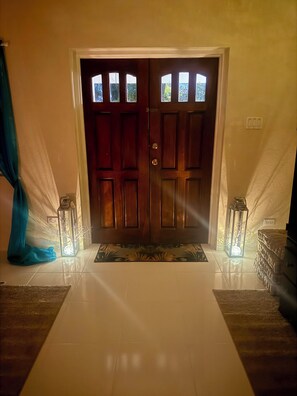 Une porte à deux battants en bois, ornée de panneaux de verre, flanquée de deux lanternes de chaque côté, qui donne sur un hall d’entrée carrelé.