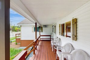 Une véranda couverte avec une terrasse en bois, des chaises blanches et un petit bâtiment en arrière-plan.