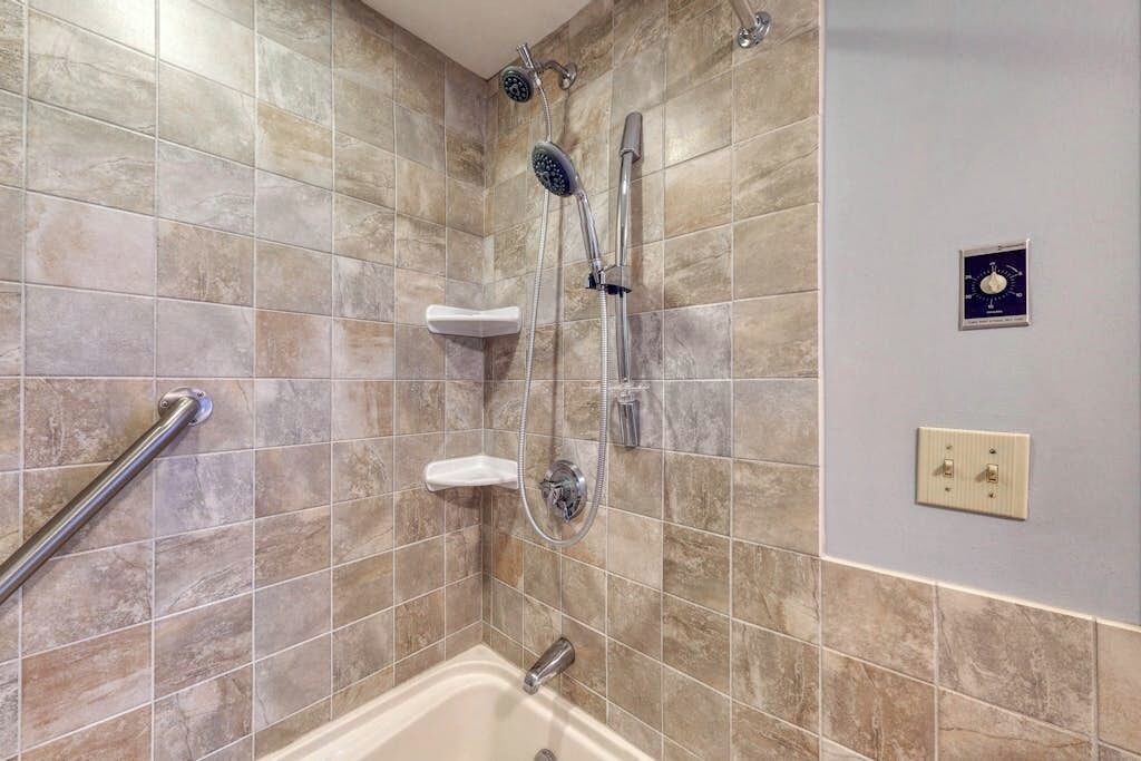 A bathroom with a shower, a handrail, a white pedestal sink, and a wall-mounted thermostat.