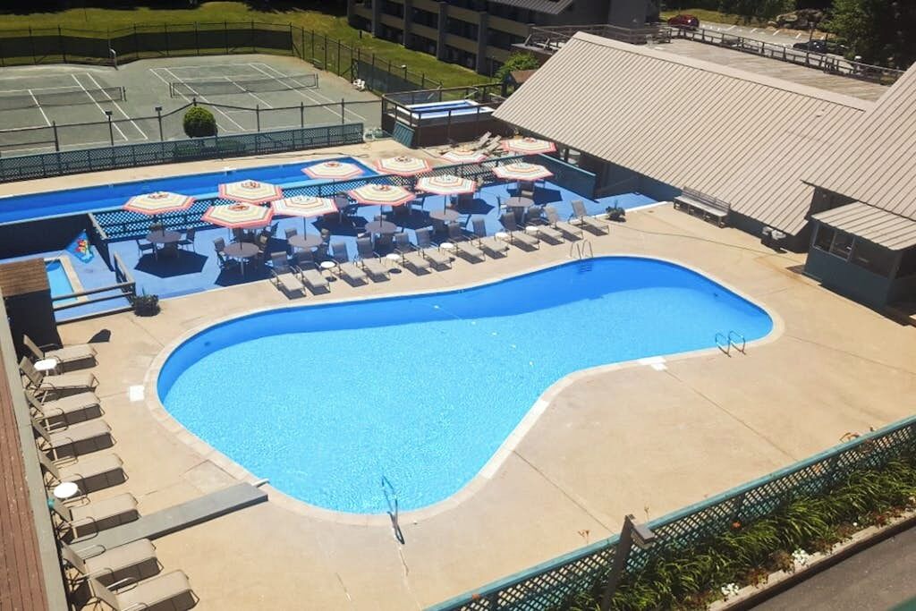 A swimming pool with sun loungers and umbrellas, surrounded by a fence and buildings.