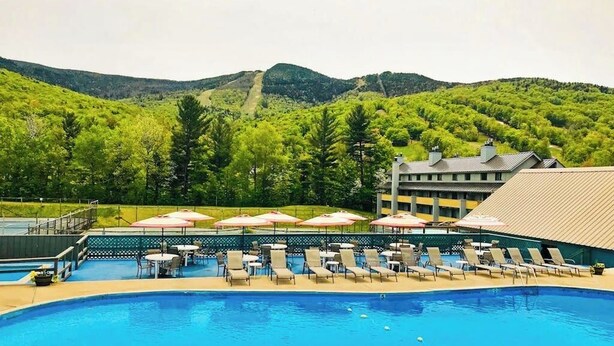 A swimming pool with lounge chairs and umbrellas, surrounded by a fence, with a mountainous backdrop.