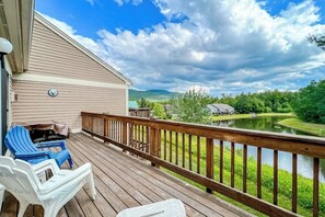 A wooden deck with chairs, a view of a lake, and a distant house. - Comfy condo with spacious deck, fast WiFi, fireplace, indoor & outdoor pools (Lincoln)
