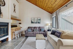 Ein Wohnzimmer mit Sofa, Couchtisch, Kamin, Küchenzeile und einem fensterbepflanzten Fenster.