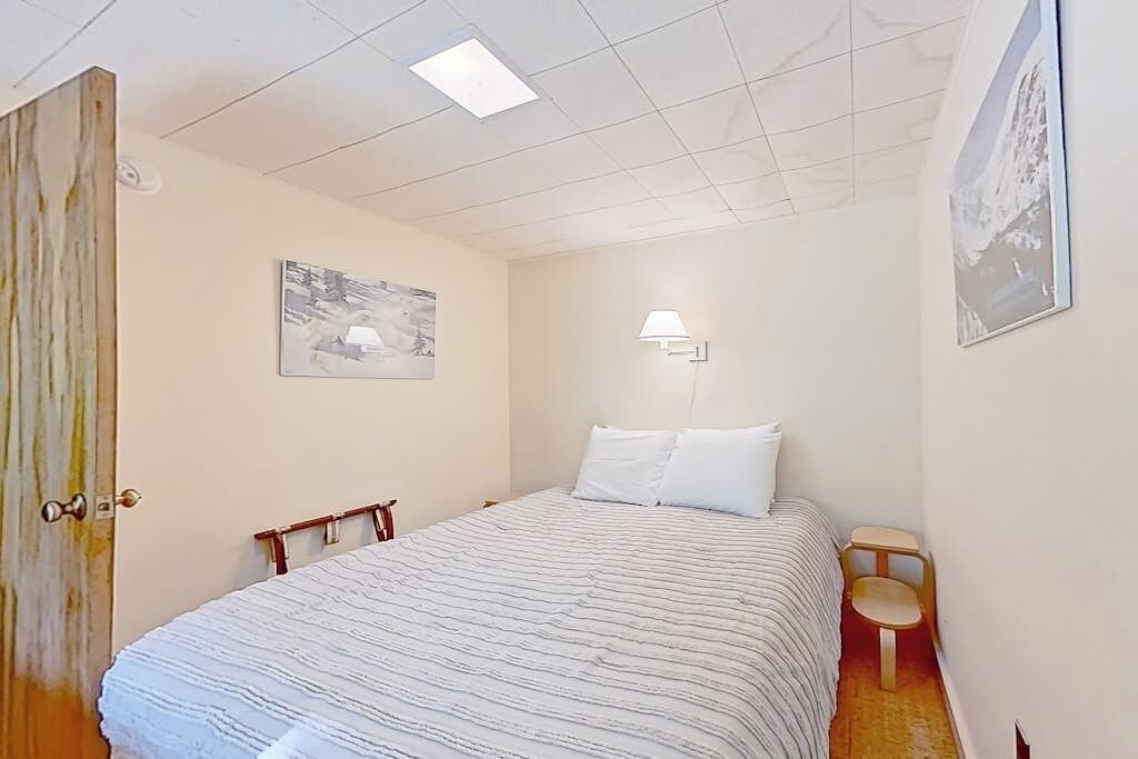 A bedroom with a bed, a nightstand, a chair, a wall-mounted lamp, and a door.