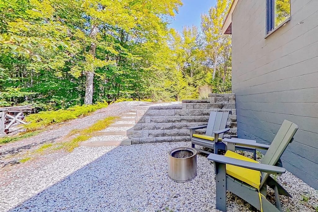 A gravel patio with Adirondack chairs, a metal fire pit, and a stone staircase leading to a building.