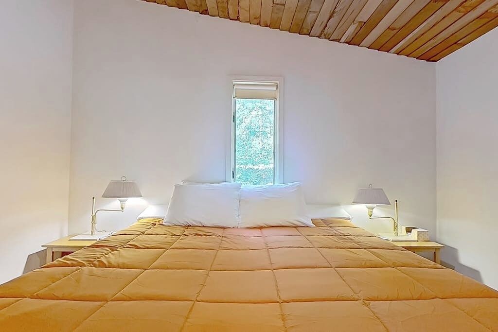 A bedroom with a bed, two bedside tables, lamps, a window, and a wooden ceiling.