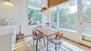 Un espace repas avec une table et des chaises en bois, de grandes fenêtres et une vue sur la verdure environnante.