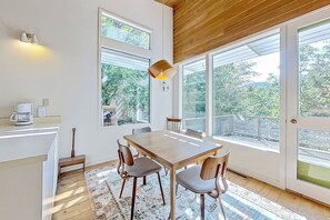 A dining area with a wooden table and chairs, large windows, and a view of greenery outside.