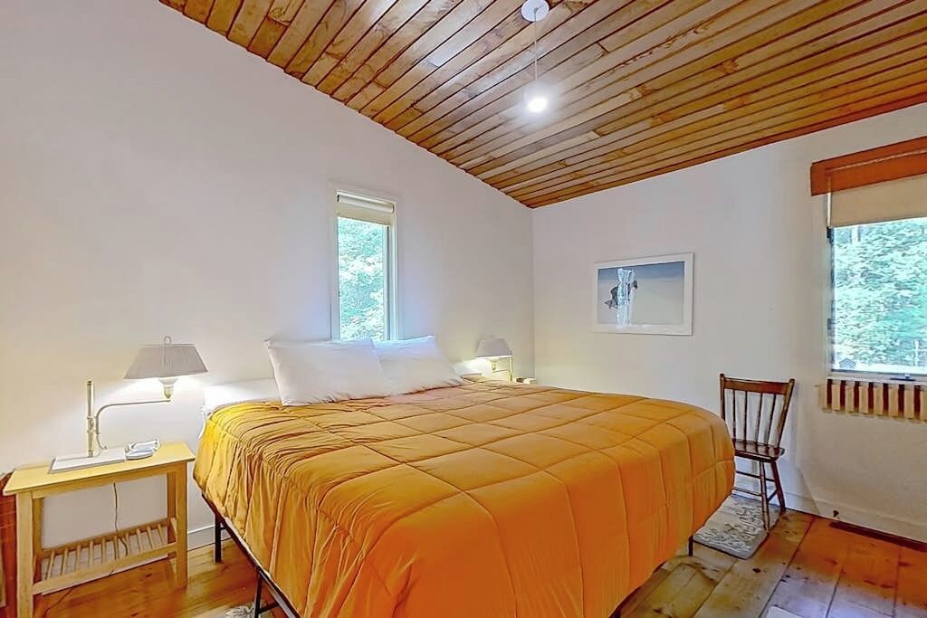 A bedroom with a wooden ceiling, a bed with an orange bedspread, a nightstand with a lamp, and a chair.