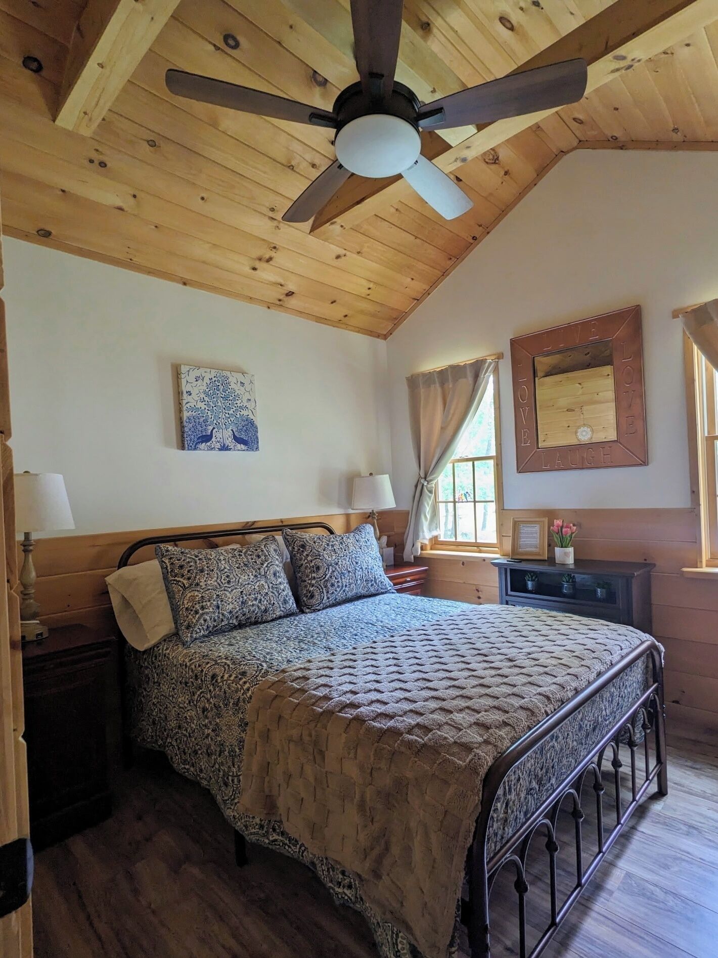 Une chambre avec un plafond en bois, un lit avec une literie à motifs, un ventilateur de plafond, un miroir fixé au mur et une fenêtre avec des rideaux.