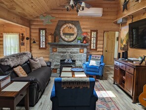 Interior de una cabaña de madera con chimenea de piedra, muebles de cuero y televisor.