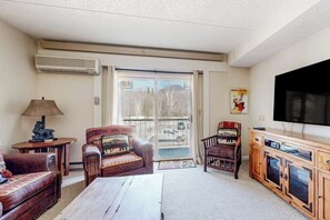 Ein Wohnzimmer mit Sofa, Ohrensessel, Couchtisch, Fernseher und einer Schiebetür zu einem Balkon.