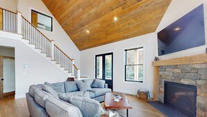 Ein geräumiges Wohnzimmer mit einem großen, grauen Ecksofa, einem Holztisch, einem Steinkamin und einer Treppe, die zu einer oberen Ebene führt.