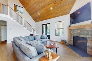 Ein geräumiges Wohnzimmer mit einem großen, grauen Ecksofa, einem Holztisch, einem Steinkamin und einer Treppe, die zu einer oberen Ebene führt.