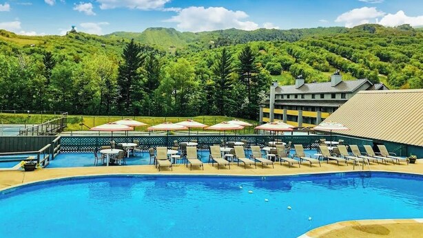 A swimming pool with lounge chairs and umbrellas, surrounded by a building with a view of green hills. - Spectacular Mountain View Condo: Aquatic Facility & Ski Shuttle (Lincoln)