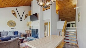 A modern living room with a sofa, coffee table, and a wooden dining table. There is a staircase with a wooden railing and a TV mounted on the wall. - Spectacular Mountain View Condo: Aquatic Facility & Ski Shuttle (Lincoln)