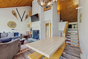 A modern living room with a sofa, coffee table, and a wooden dining table. There is a staircase with a wooden railing and a TV mounted on the wall. - Spectacular Mountain View Condo: Aquatic Facility & Ski Shuttle (Lincoln)
