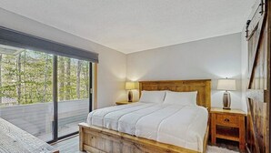 A bedroom with a wooden bed, two bedside tables with lamps, a large window with a view of trees, and a white furry rug. - Spectacular Mountain View Condo: Aquatic Facility & Ski Shuttle (Lincoln)