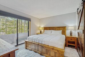 A bedroom with a wooden bed, two bedside tables with lamps, a large window with a view of trees, and a white furry rug. - Spectacular Mountain View Condo: Aquatic Facility & Ski Shuttle (Lincoln)