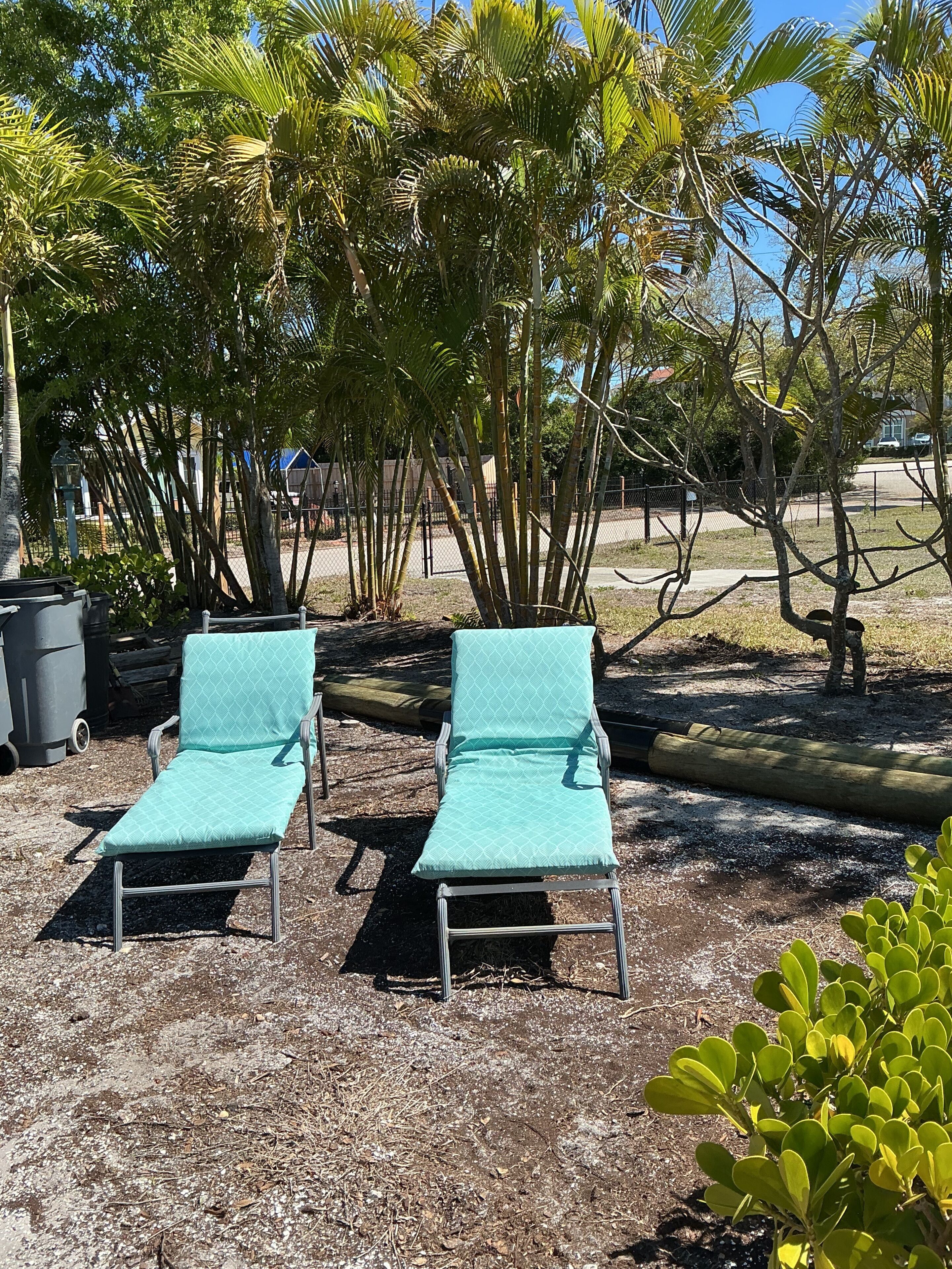 Two lounge chairs with metal frames and light blue cushions, positioned on a gravel surface with surrounding palm trees.