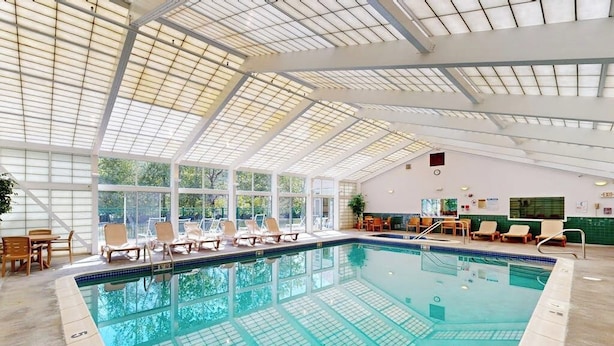 Une piscine intérieure avec des parois en verre transparent, des chaises longues et un plafond haut doté de lucarnes.