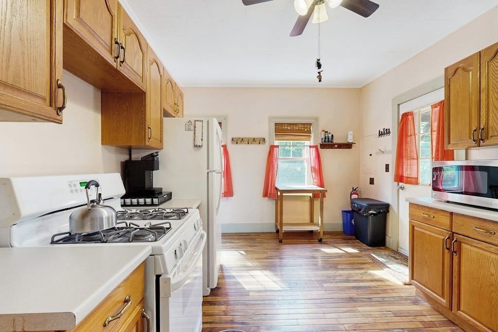 A kitchen with wooden cabinets, a stove, a refrigerator, a ceiling fan, and a window with curtains.