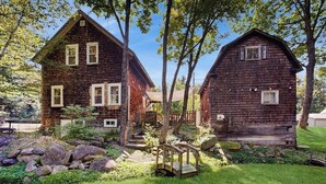 Une cabane en bois rustique, avec un pont en bois, entourée d’un jardin bien entretenu et d’arbres.