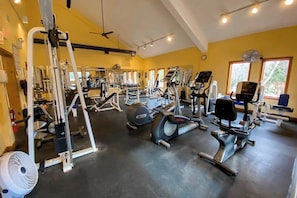 Ein Fitnessraum mit unterschiedlichen Trainingsgeräten, einem Deckenventilator und großen Fenstern.