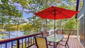 Una terraza con mesa y sillas de vidrio, un paraguas rojo y vistas a un lago.