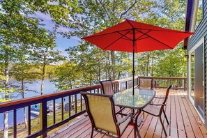 A deck with a glass table and chairs, a red umbrella, and a view of a lake. - Stunning lakefront home with private dock, deck, firepit, and lake view (Wakefield)