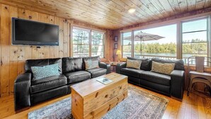 A cabin interior with two black leather sofas, a wooden coffee table, a mounted TV, and large windows with a view of trees.