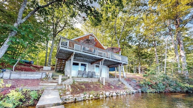 A two-story cabin with a balcony, surrounded by a pond and trees.