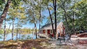 A cabin by a lake with a deck, chairs, and a small table.
