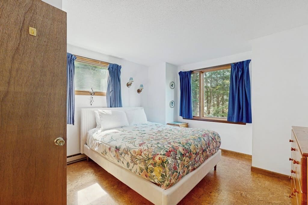 A bedroom with a bed, two windows with curtains, a wooden door, and a dresser.