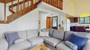 Una sala de estar con un sofá seccional gris, una escalera de madera y una cocina visible a través de un vano abierto.