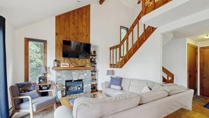 Una sala de estar con chimenea de piedra, un sofá, un sillón, un televisor montado en la pared y una escalera de madera.