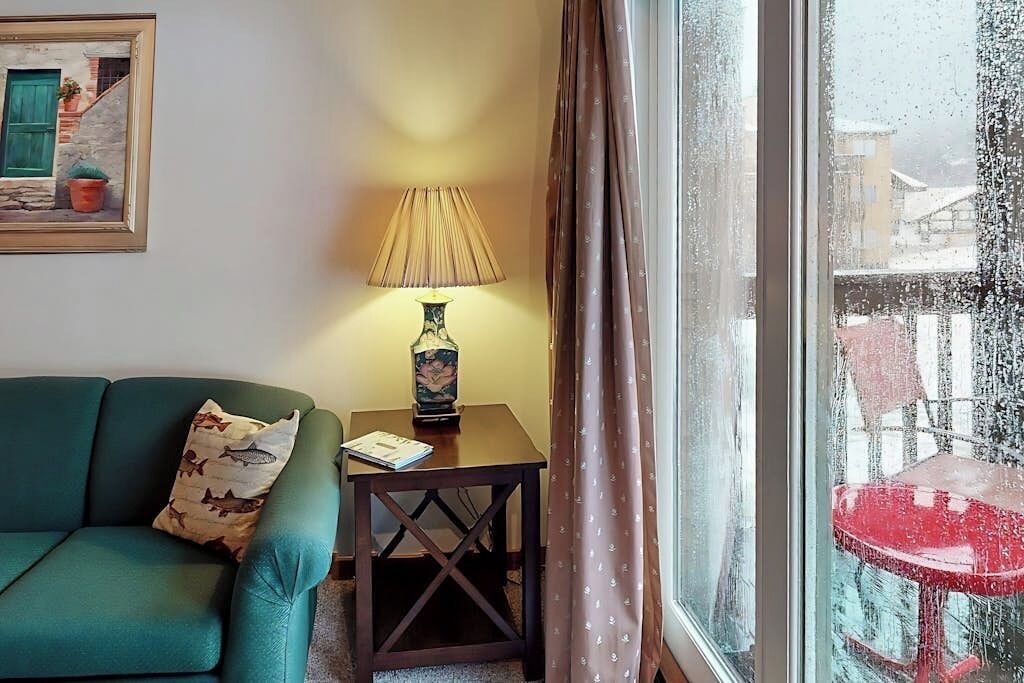 A room with a green sofa, a small table with a lamp, and a window with a view of buildings.