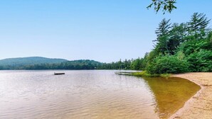 A calm lake with a sandy shore, surrounded by dense forest and a hill in the background. - Open concept home close to the beach with gas fireplace and deck (Madison)