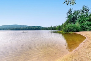 A calm lake with a sandy shore, surrounded by dense forest and a hill in the background.