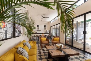Un salon moderne avec un sol à carreaux, un canapé jaune, des fauteuils en osier et un coin repas comprenant une table et des chaises.