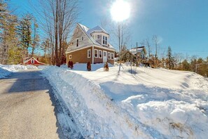 A two-story house with a sloped roof, a front porch, and a snow-covered yard. - 2-story dog-friendly home in downtown with grill, mtn views, streaming, & porch (North Conway)