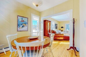 A dining area with a wooden table and white chairs, a painting of a ski lift, and a view into a living room with a red sofa. - 2-story dog-friendly home in downtown with grill, mtn views, streaming, & porch (North Conway)
