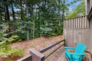 Eine Holzterrasse mit einem türkisblauen Adirondack-Sessel, umgeben von bewaldetem Gelände mit hohen Bäumen und einem Holzzaun.